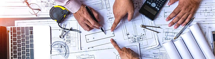 Top-down image of hands of people working on blueprints, with laptop, drafting instruments